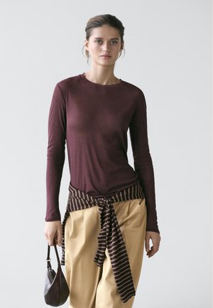 Kvinde iført en bordeaux langærmet bluse, beige bukser med vide ben, stribet sweater bundet om taljen, og hun holder en lille mørk håndtaske.