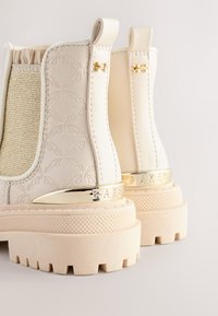 Bottes blanches à la cheville avec un motif texturé, des panneaux latéraux élastiques, des accents dorés et une semelle beige épaisse avec une prise texturée.