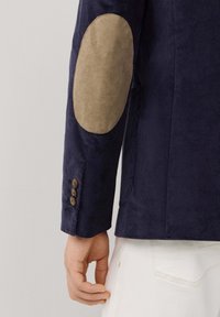 Chaqueta de pana azul marino con parches de ante en color beige en los codos, puños de tres botones y un corte entallado. Tejido texturizado con un patrón de costilla vertical.