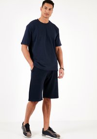 Tricou navy blue cu mâneci scurte și tivuri rulate, asociat cu pantaloni scurți până la genunchi asortați. Încălțămintea are șireturi negre cu accente gri.