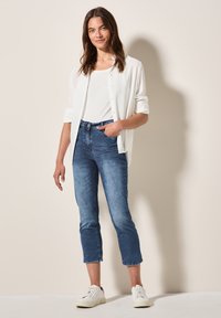 Chemise blanche à boutons, débardeur blanc ajusté, jeans en denim bleu clair, longueur raccourcie avec ourlet brut, et baskets blanches. Fond neutre.