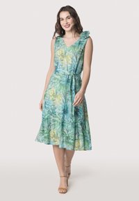 Robe florale dans des tons verts et jaunes, avec un décolleté en V, sans manches, une taille ajustée avec une ceinture à nouer, et une jupe fluide.