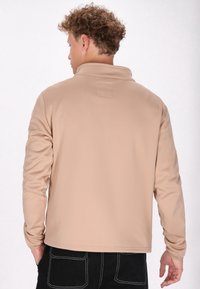 Beige jack met een kraagontwerp, een gladde textuur, lange mouwen en een klein vierkant patch op de rug. Draagt over zwarte broek.