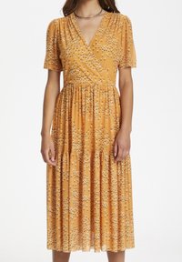 Mujer vestida con un vestido midi amarillo mostaza con pequeño estampado floral blanco, de manga corta, escote en V y cintura fruncida.