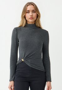 Vrouw met blond haar die een aansluitend donkergrijs shirt met lange mouwen en een gouden ringdetail op de taille draagt, samen met zwarte broek.