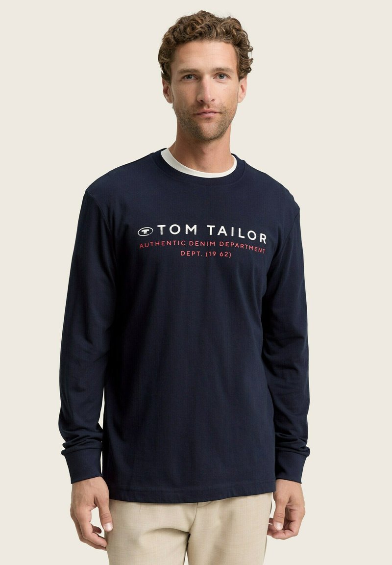 Camiseta de manga larga azul marino con cuello redondo, texto impreso en blanco y rojo, y un corte relajado. La tela es suave y ligeramente elástica.