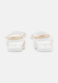 Białe sandały o gładkiej teksturze, z beżowym akcentem na obcasie, regulowanym paskiem z tyłu oraz teksturowaną gumową podeszwą.