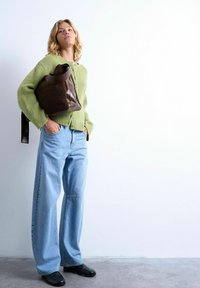 Jeune femme portant un pull vert en tricot et un jean à jambes larges, avec un grand sac en cuir marron et en peau lainée, posant devant un mur blanc.