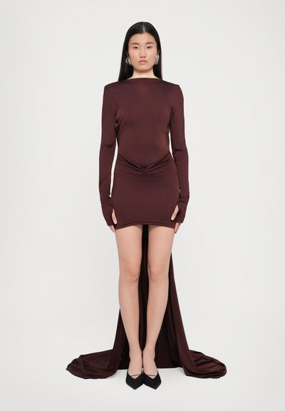 Robe bordeaux à manches longues avec un corsage ajusté, des drapés à la taille et un ourlet arrière plus long. Tissu lisse ; associée à des chaussures noires.