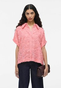 Roze shirt met korte mouwen en een textuur met rafelige details en een kraag. Gecombineerd met donkere jeans en een bruine clutch.