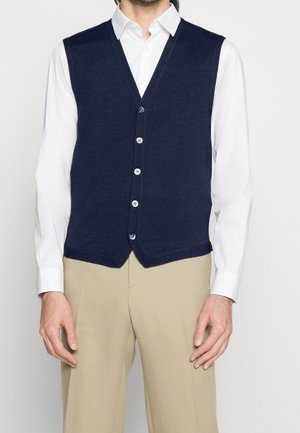 Mann trägt eine marineblaue, geknöpfte Weste über einem weißen Langarmhemd und beigen Hosen, steht vor einem schlichten weißen Hintergrund.