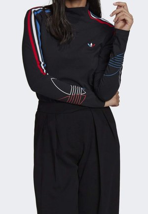 Femme portant un haut noir à manches longues avec des rayures rouges, bleues et blanches sur les manches et un pantalon large noir assorti, debout les bras croisés.