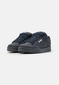 Black sneakers with a textured finish, featuring a white logo, contrasting stitching, and thick black laces. Low-top design and rubber sole.