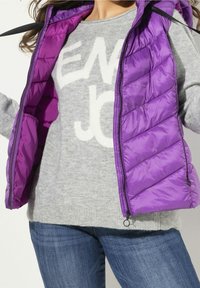 Fialová prošívaná vesta typu puffer, nošená přes světle šedý svetr s bílou grafikou. Outfit doplňují tmavě modré džíny.