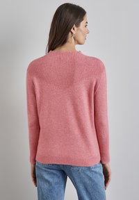 Różowy sweter wykonany na drutach z żebrowanym dekoltem i detalem na plecach. Posiada długie rękawy oraz prosty dół, zestawiony z jasnoniebieskimi dżinsami.