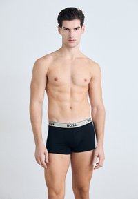 Schwarze Boxer-Shorts mit einem goldakzentuierten Bund, der den Text "BOSS" zeigt. Der Stoff wirkt glatt, das figurbetonte Design betont die Silhouette.