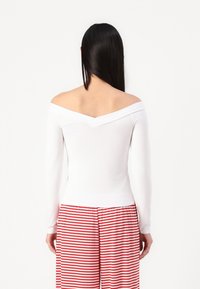 White long-sleeve off-the-shoulder top, featuring a V-shaped back neckline, paired with red and white striped pants. Smooth fabric texture.