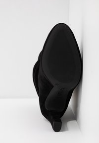 Bottines noires en suède à talons hauts, à bout pointu, posées en angle contre un mur blanc, montrant la semelle texturée et la marque de taille 37.