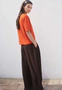 Orange Kurzarmoberteil kombiniert mit lockeren braunen weiten Hosen. Glatter Stoff, entspannter Schnitt und seitliche Taschen sind in den Hosen sichtbar.