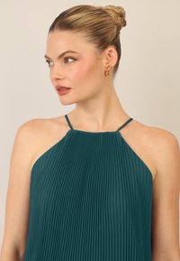 Turkusowa sukienka halter z fałdami i cienkimi ramiączkami, prezentująca fakturowany materiał. Modelka nosi złote kolczyki w kształcie kół. Prosty, elegancki design.