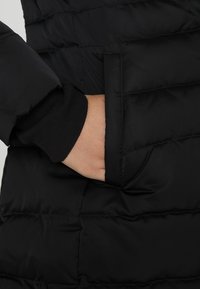 Musta puffer-tikkitakki, jossa on tikattu pinta ja sivutasku; ranteen resorissa on joustavaa kangasta mukavuutta ja tiukkaa istuvuutta varten.