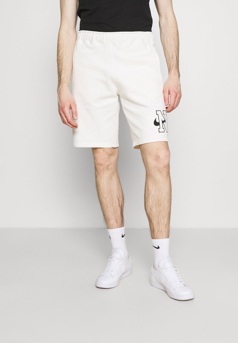 Beige shorts med resår i midjan och svart logotypdetalj, kombinerade med vita sneakers och strumpor. Enkel, rakt skuren design, lämplig för vardagsbruk.
