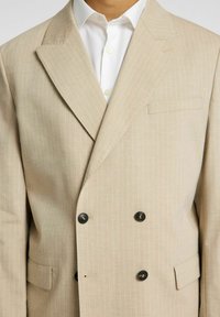 Beige dvouřadý blazer s tenkými vertikálními pruhy, s vroubkovanými klopami, dvěma předními kapsami a černými knoflíky. Hladká textilie.