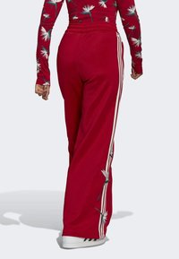 Red track pants with a relaxed fit, featuring white side stripes and floral embroidery along the left leg, made from soft fabric.