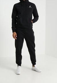 Černá mikina s kapucí a klokankou, spárovaná s odpovídajícími tepláky. Obsahuje bílé logo na hrudi a na kalhotách. Bílé tenisky.