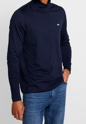 Navyblaues Langarm-Turtleneck-Shirt aus weichem Stoff, mit einem kleinen Logo auf der Brust, kombiniert mit blauen Denim-Jeans.