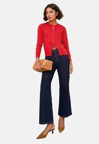 Kvinna som bär en röd knappad kofta, mörka utsvängda jeans, bruna slingback-klackar och håller en beigefärgad läderclutch.