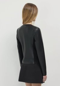 Donna con lunghi capelli castano chiaro, che indossa un top aderente in pelle nera a maniche lunghe e una gonna nera, vista di spalle su uno sfondo neutro.