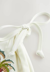 White, knitted fabric top with a knotted strap. Features colorful sequined palm leaf embroidery in green, gold, and bronze.