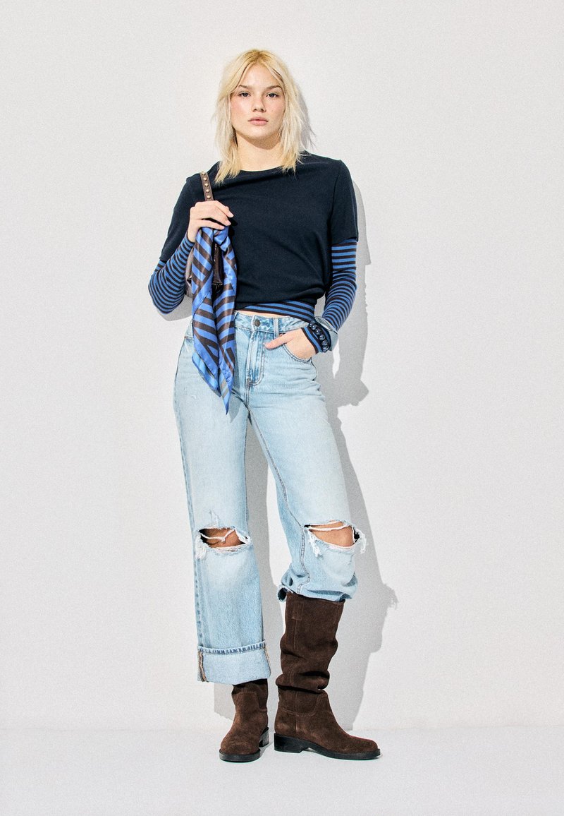 Mladi žena plave kose koja nosi višeslojnu tamnoplavu i plavu prugastu dugih rukava, crnu majicu, izrezane svijetle traperice s obrubom i smeđe brušene čizme.