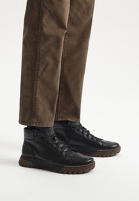 Czarne skórzane sneakersy typu high-top o teksturowanym wykończeniu, z zawiązywaniem na sznurówki i brązową gumową podeszwą. Noszone z brązowymi spodniami, eksponując wysokość do kostki.