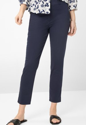 Frau trägt marineblaue, eng anliegende Hose, langärmliges Oberteil mit Blumenmuster und schwarze flache Sandalen, steht vor weißem Hintergrund.