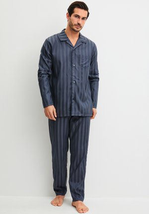 Mann steht barfuß und trägt ein dunkelblau gestreiftes, langärmliges Pyjamaoberteil mit Knöpfen und passende Hose vor einem einfarbigen weißen Hintergrund.