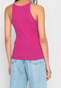 Fitted, ribbed tank top in bright pink with a high neckline and racerback design. Paired with light blue, high-waisted denim jeans.