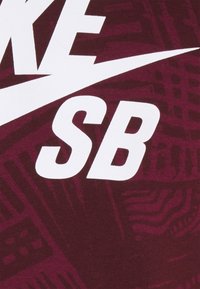 Burgundy-kangas, jossa on suurta valkoista "NIKE"-tekstiä ja "SB"-logo, taustalla teksturoitu kuvio. Silkkinen puuvillamateriaali.