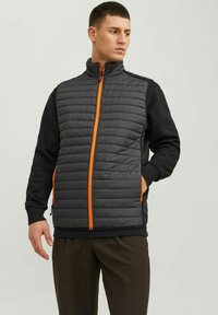 Gepolsterte Weste mit einer gesteppten grauen Vorderseite und schwarzen Ärmeln, ausgestattet mit einem orangefarbenen Reißverschluss und Taschen, die ein Mischdesign aus verschiedenen Texturen bieten.