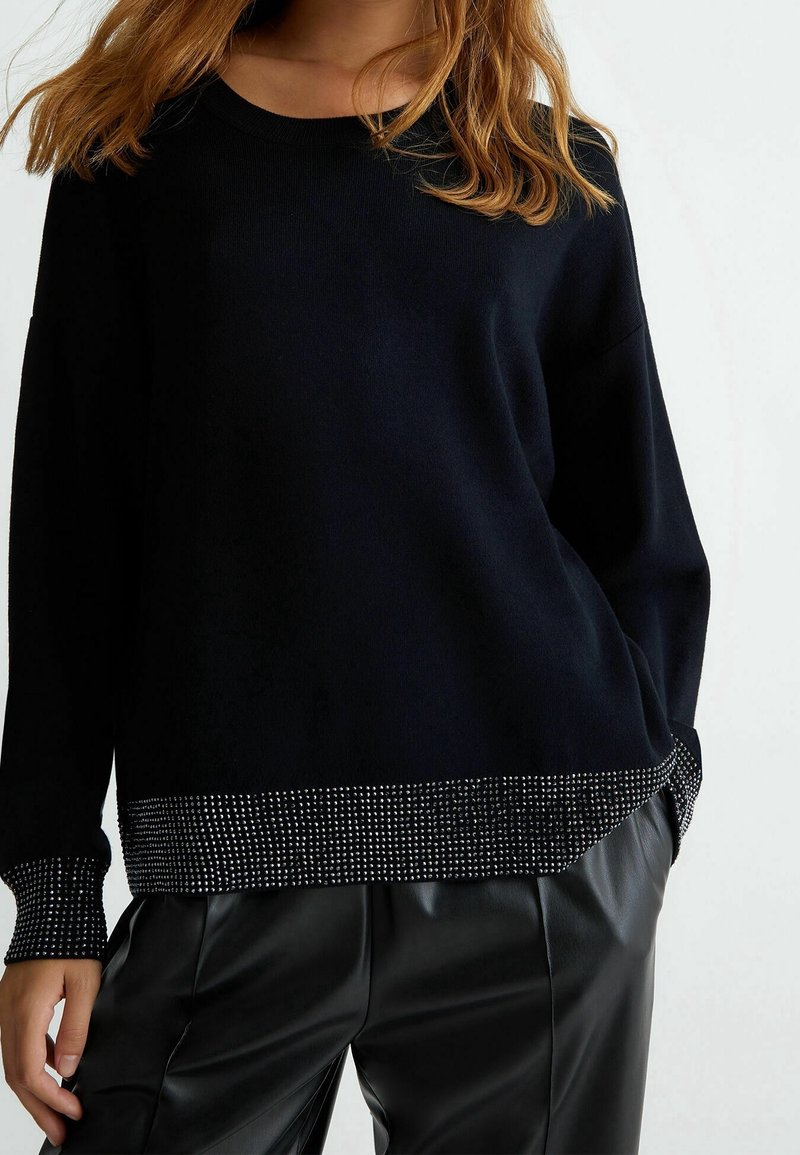 Femme portant un pull noir à manches longues avec de petits clous argentés aux poignets et à l'ourlet, associé à un pantalon en cuir noir, main dans la poche.