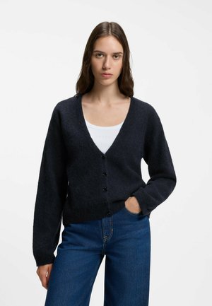 Jeune femme aux longs cheveux bruns portant un cardigan noir boutonné par-dessus un top blanc et un jean bleu, debout devant un fond blanc uni.