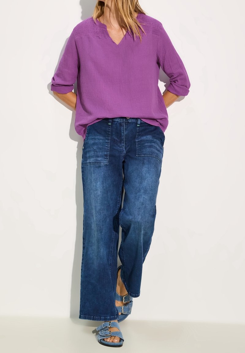 Blouse violette décolletée en V, ample avec des manches 3/4, associée à un jean en denim sombre à jambes larges. Accentuée par des sandales à double boucle.
