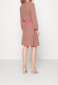 Robe en tissu tissé avec un motif en grille rose et beige, dotée de longues manches et d'une taille ceinturée, terminée par un ourlet contrastant rose.