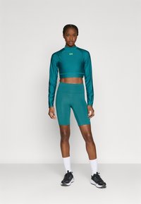 Μπλε-πράσινο cropped τοπ με μακρύ μανίκι και ταιριαστά σορτς ποδηλασίας με ψηλή μέση, κατασκευασμένα από λείο ύφασμα. Συνδυασμένα με μαύρα αθλητικά παπούτσια και λευκές κάλτσες.