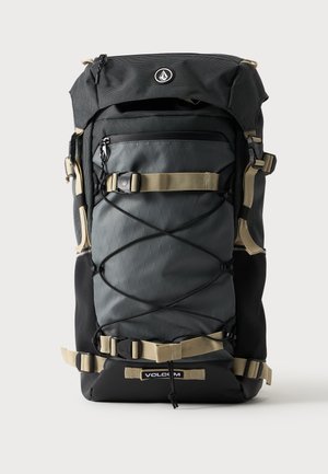 Grand sac à dos noir et gris avec des bretelles beige, un cordon élastique à l'avant, une poche zippée et le logo Volcom en bas au centre.