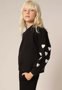 Zwarte sweatshirt met gestructureerde witte hartpatronen op de mouwen. Heeft een relaxte pasvorm en een geribde onderrand, gemaakt van zachte, gladde stof.