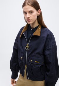 Νεαρή γυναίκα φορώντας σκούρο μπλε μπουφάν Barbour με καφέ γιακά από κορδόνι και μπεζ παντελόνι πάνω σε απλό ανοιχτόφωτο φόντο.