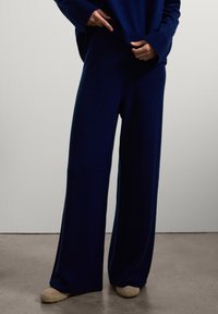 Personne portant un pantalon en tricot large bleu marine et un pull assorti, se tenant sur un sol gris contre un mur blanc.