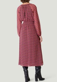 Robe midi à manches longues en tissu transparent avec un motif géométrique rose et noir, taille smockée et col rond. Portée avec des bottes noires.